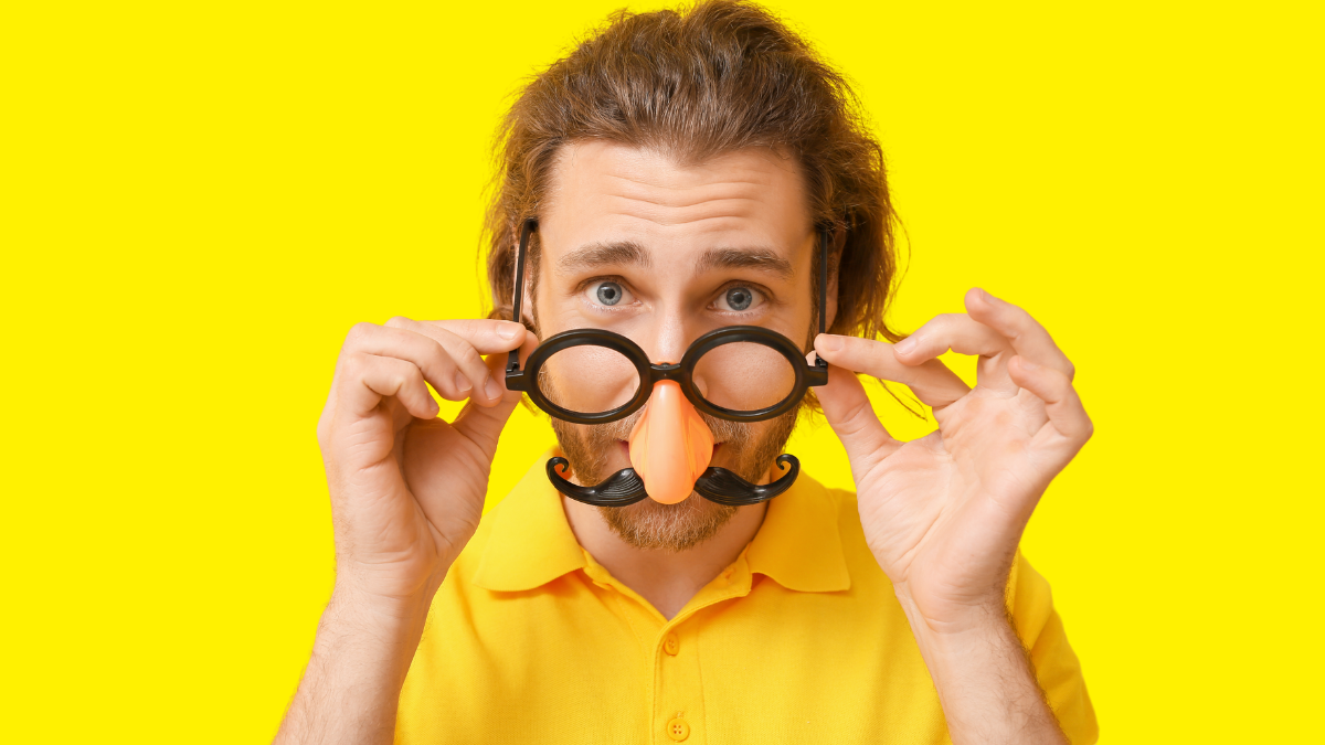 A man holding up fake glasses and a moustache disguise against a bright yellow background, symbolizing how fake candidates can hide behind AI tools and deceptive identities during hiring.